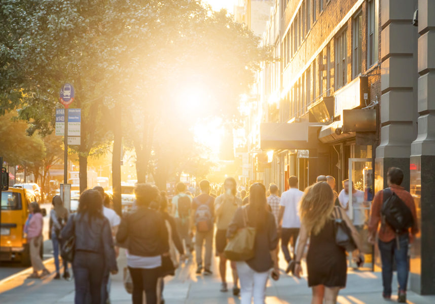 Man stands in the middle of a busy sidewalk looking at his cell phone while crowds of people walk around on 14th Street in Manhattan, New York City with the glow of sunlight in the background.